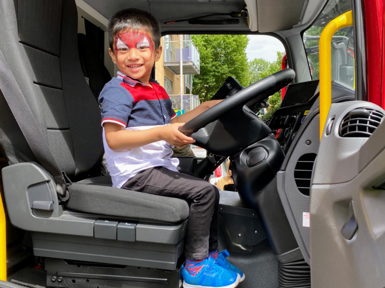 A young boy with spider man facepaints smiles happily as he sits in the driver seat of a fire engine at a Summer of Wellbeing Festival