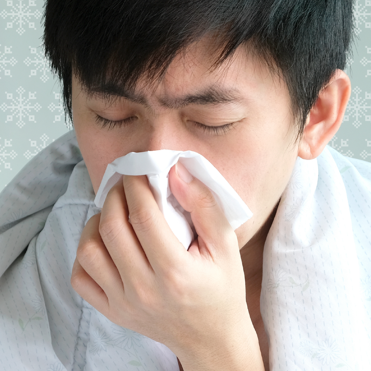 A young man is bundled up in a duvet, blowing his nose and looking ill.