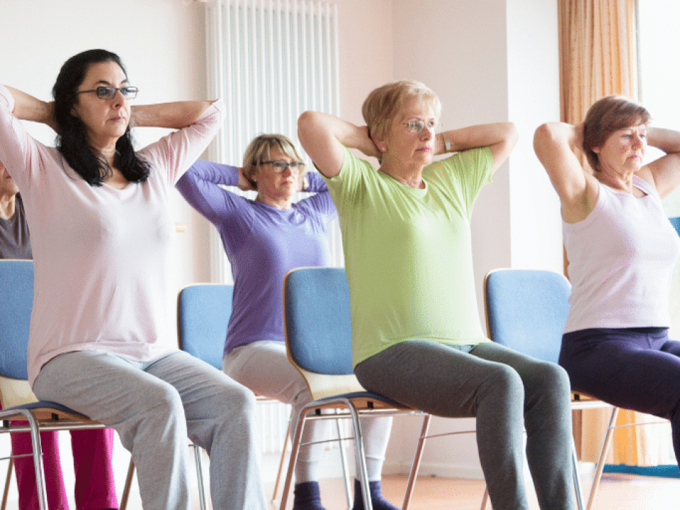 People at a seated exercise class, gently stretching with their hands behind their heads.