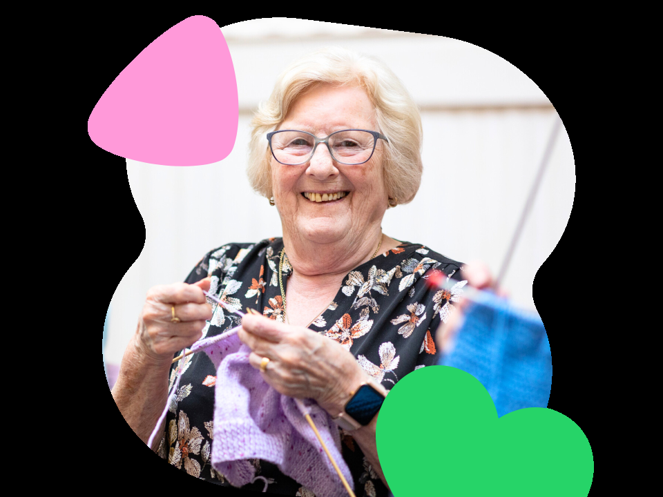 An older woman is chatting and laughing as she knits at a knit and natter session